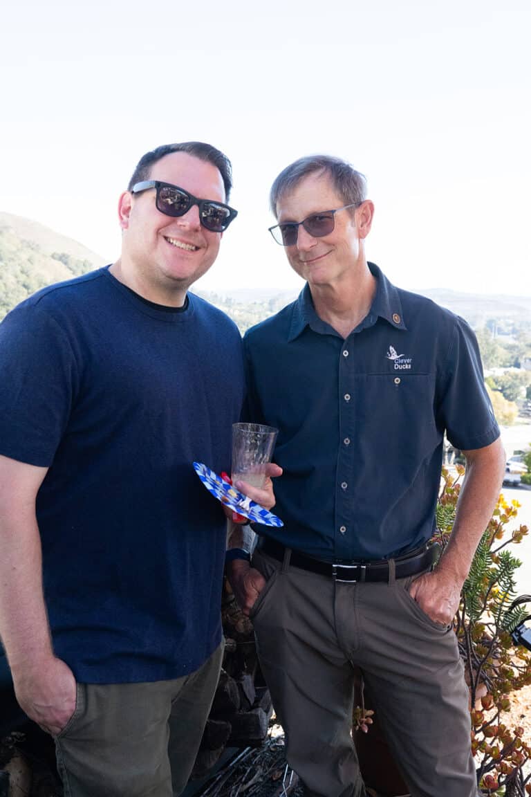 Two team members in sunglasses smiling on a hillside patio, one in a Clever Ducks shirt holding a drink