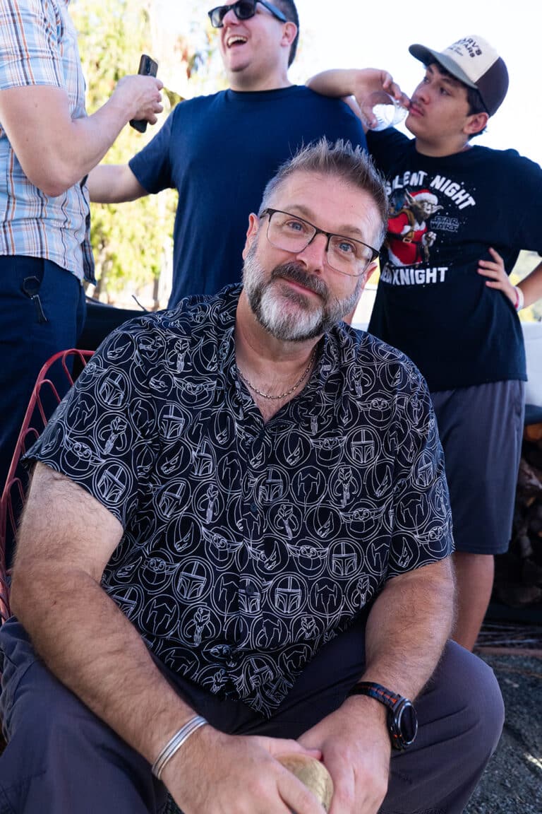 A team member in glasses and a patterned short-sleeve shirt smiling at the camera at an outdoor party, two others laughing behind him