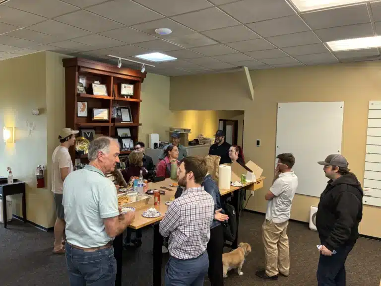 Team members standing around a table of food in the office, a small tan dog among them