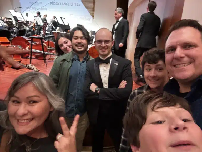 A group selfie on a concert stage with a musician in a tuxedo, orchestra instruments behind them