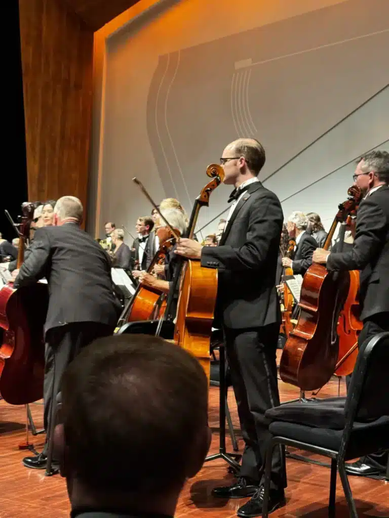 An orchestra on stage seen from the audience, a cellist in a tuxedo standing with his cello