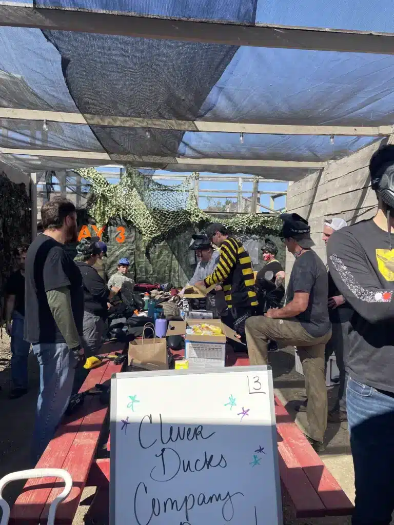 Team members in paintball masks around a picnic table under camouflage netting, a Clever Ducks Company sign in front